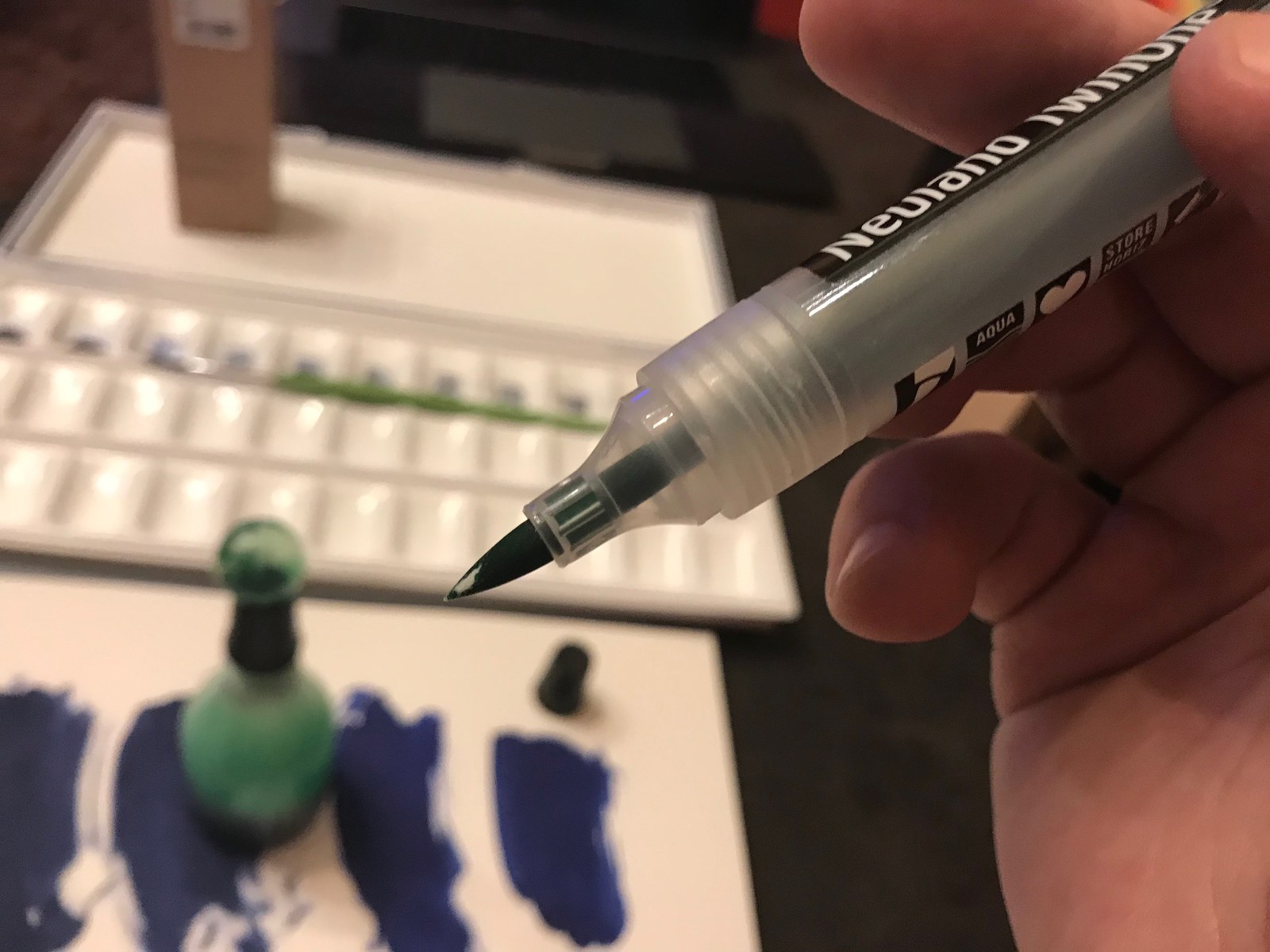 A hand holding a brush marker above refill supplies and painted color swatches.